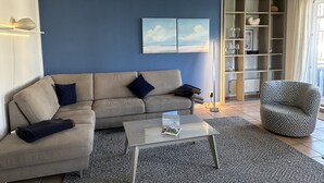 Ein Wohnzimmer mit einem beigefarbenen Sofa, einem Glastisch und einem gemusterten Sessel. An der Wand hängt ein Bild, und ein Bücherregal beherbergt Glaswaren.