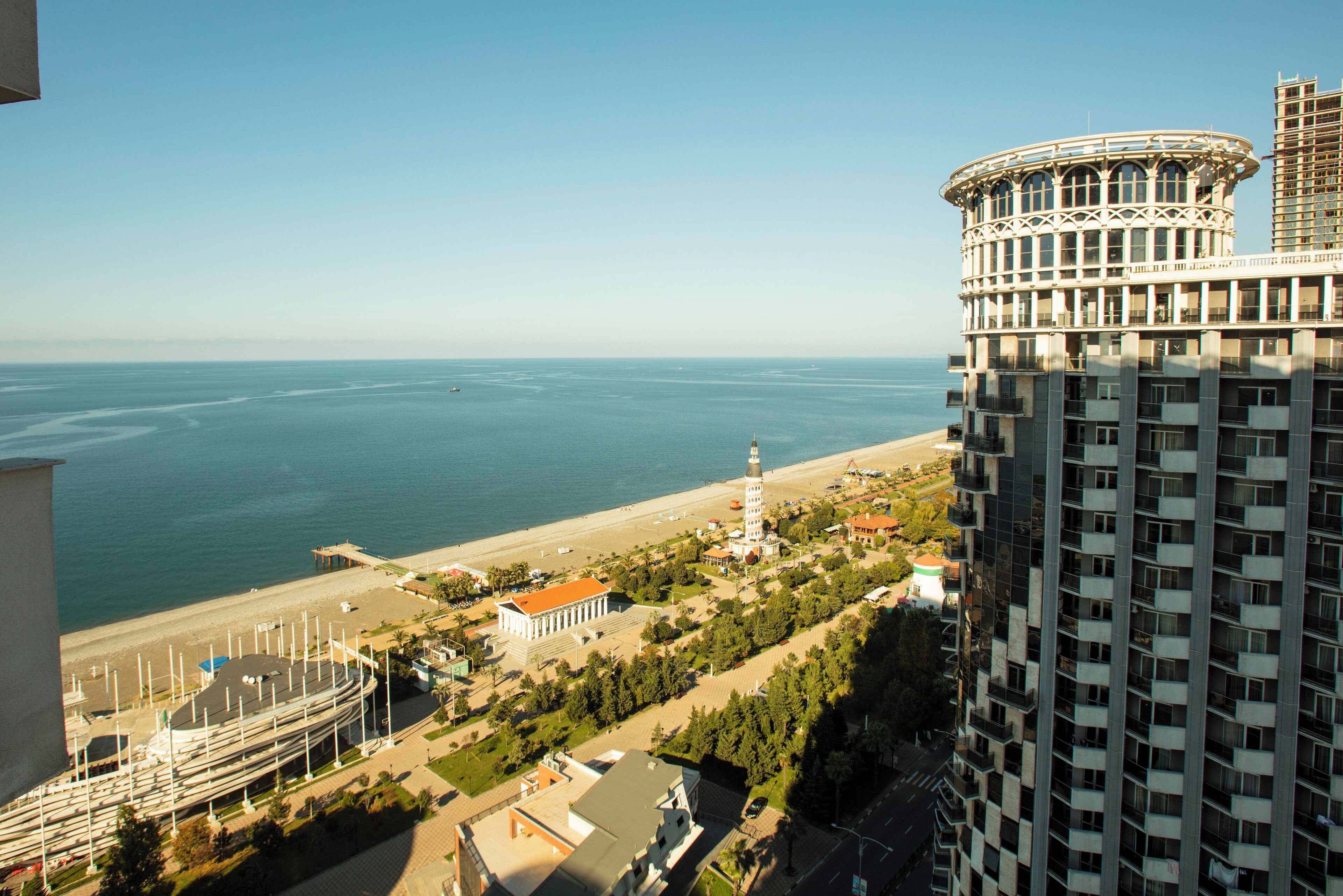 A high-rise hotel overlooking a beach with a lighthouse, a mosque, and a round building with a balcony.