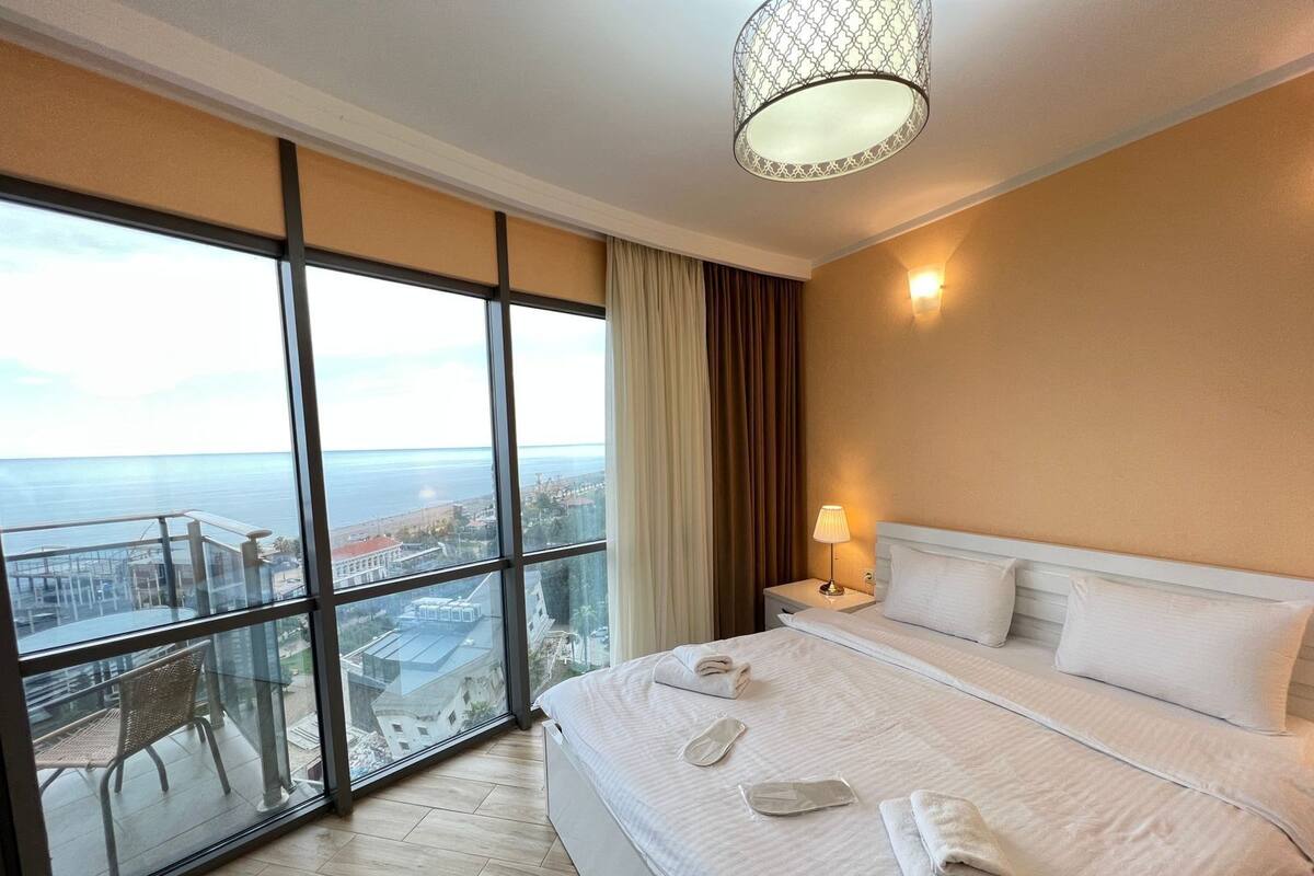 Une chambre d’hôtel dotée d’une grande fenêtre donnant sur la ville, d’un lit avec du linge de lit blanc, d’une lampe de chevet et d’une vue sur la mer.