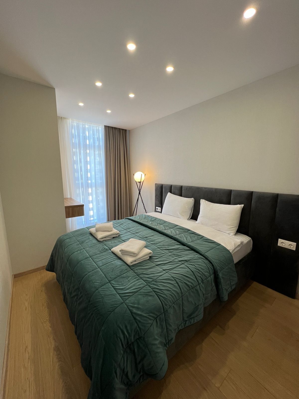 A modern hotel room with a large bed, a bedside lamp, and a view of the city through the window.