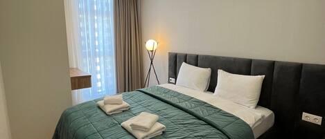 A modern hotel room with a large bed, a bedside lamp, and a view of the city through the window.