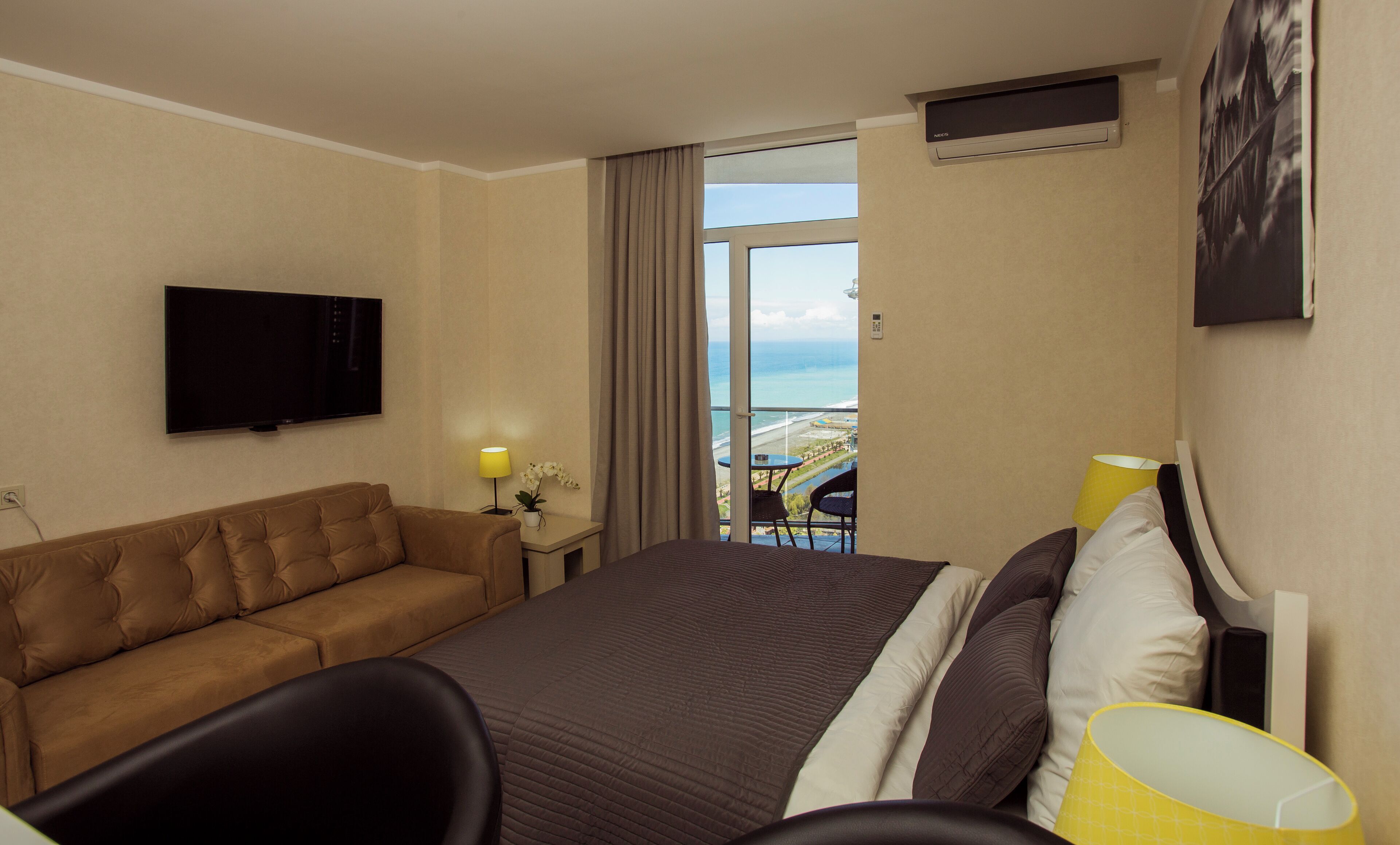 Een hotelkamer met een bed, een bank, een televisie, een raam met uitzicht op zee en een balkon.