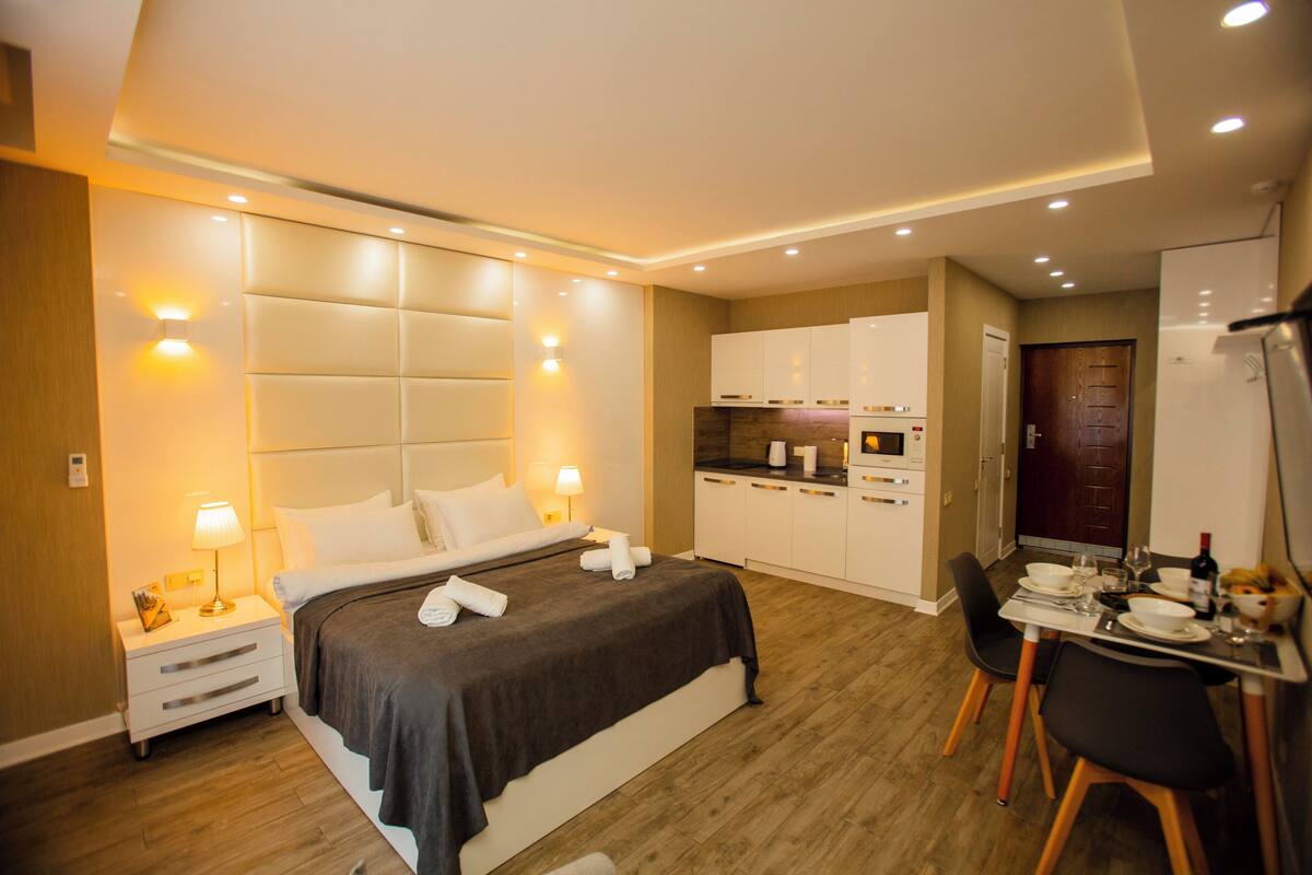 Une chambre d’hôtel moderne dotée d’un grand lit, de tables de chevet, d’un bureau avec des chaises et d’une kitchenette.