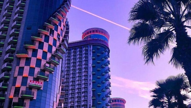 Un immeuble de grande hauteur, doté d’une façade à motifs de carreaux distinctifs et de balcons, entouré de palmiers et surplombant un ciel au coucher du soleil.