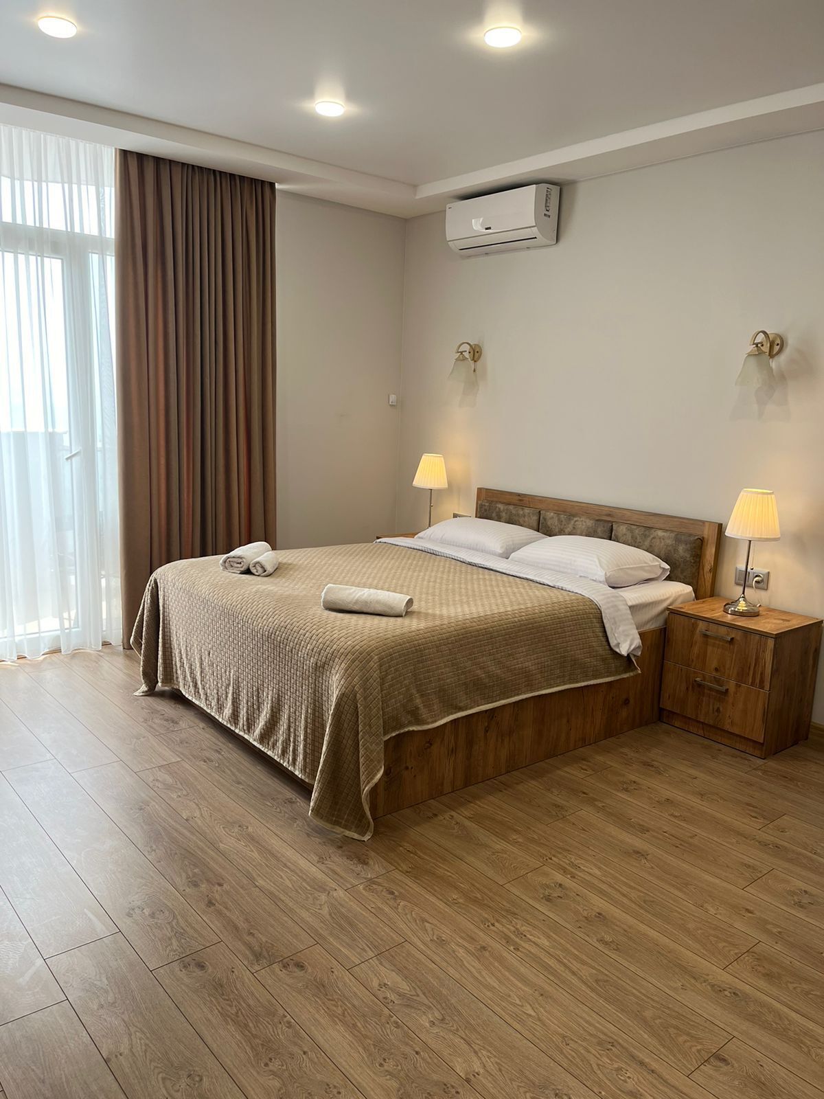 Una cama bien hecha con un cabecero de madera, dos mesitas de noche con lámparas, y un equipo de aire acondicionado en la pared.