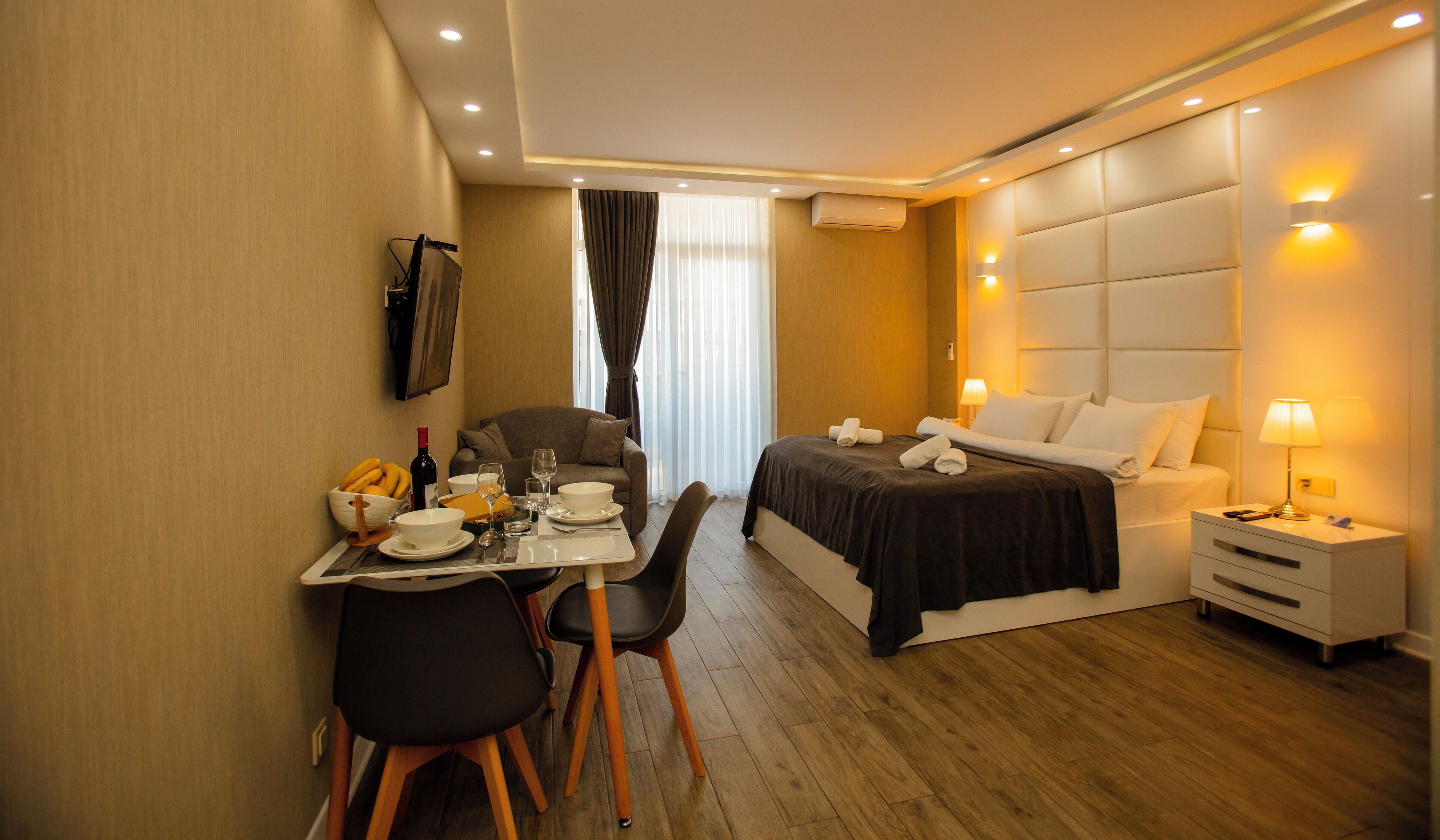 A hotel room with a dining table set for two, a bed with a headboard, bedside tables with lamps, and a flat-screen TV mounted on the wall.