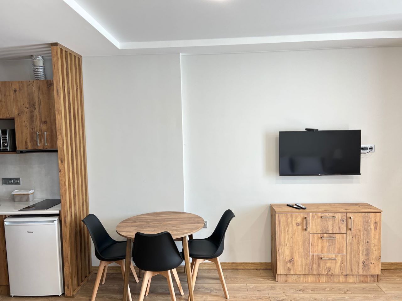 A compact living space with a kitchenette, a dining area with a round table and four chairs, a flat-screen TV mounted on the wall, and a wooden cabinet.