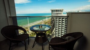 Un balcon avec des fauteuils en osier et une table en verre, d’où l’on a vue sur une plage et un grand immeuble.