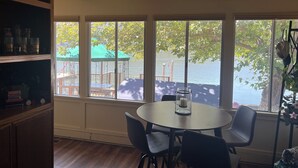 A dining area with a round table and chairs, large windows with a view of a dock and greenery, and a shelf with various items.