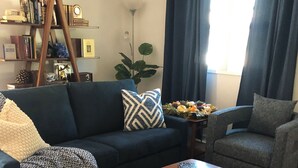 A living room with a dark blue sofa, a wooden coffee table, a patterned throw blanket, and a bookshelf with various books and decorative items.