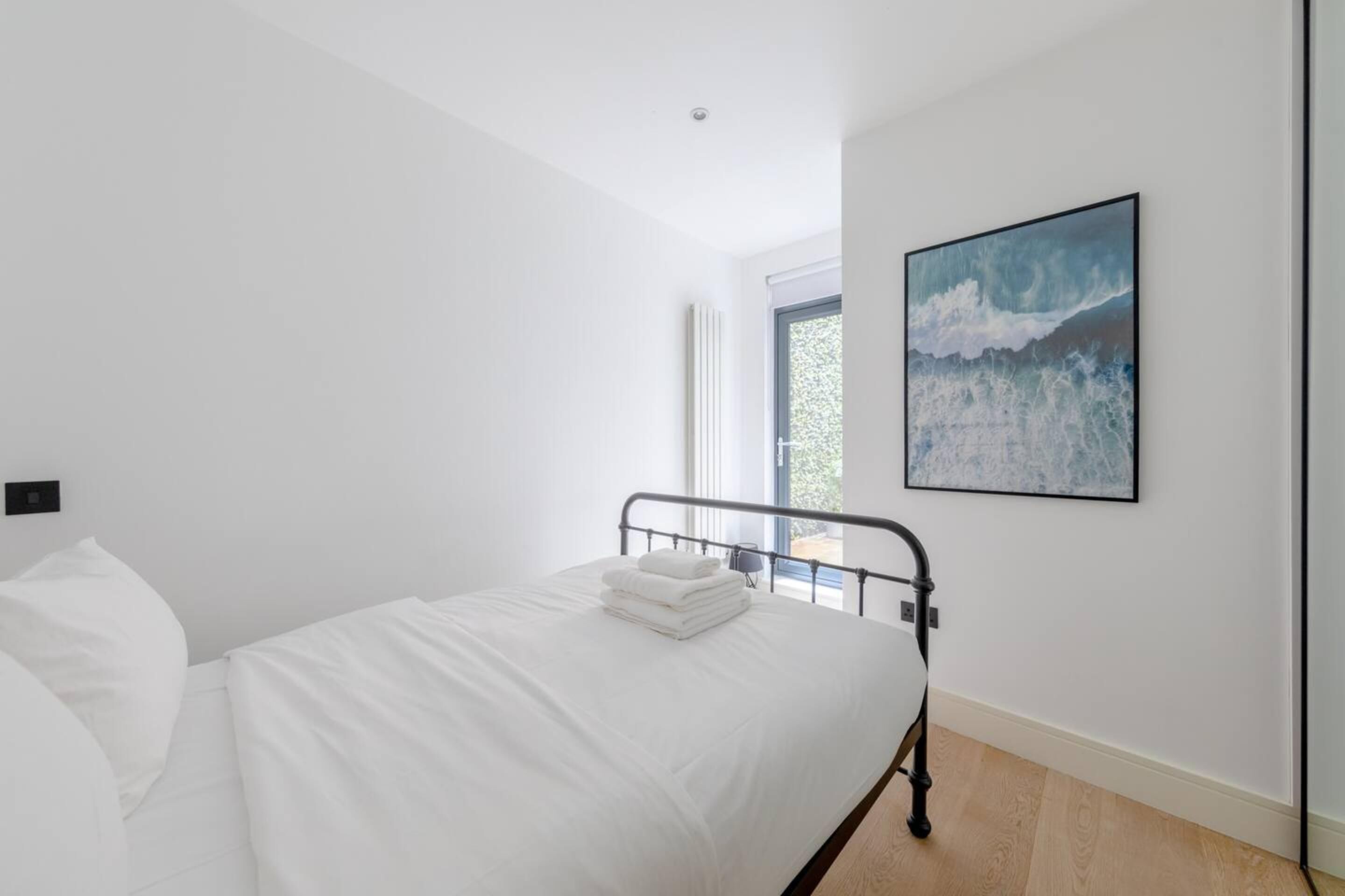Ein modernes Schlafzimmer mit einem Einzelbett, weißen Bettwäsche, einem Metall-Kopfteil, einem großen Fenster und einem gerahmten abstrakten Gemälde.