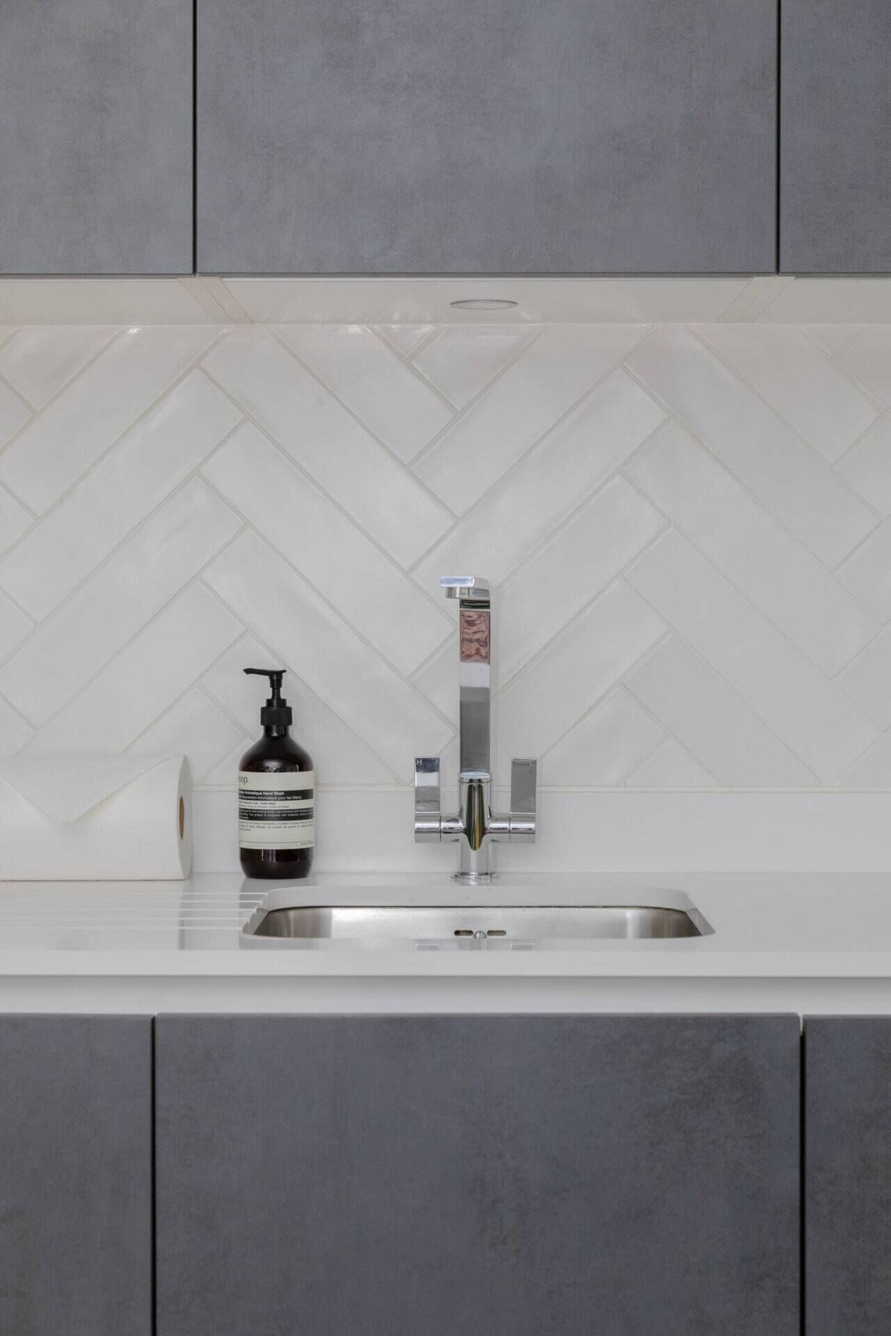 A modern kitchen with a white herringbone tile backsplash, a stainless steel sink, and a soap dispenser.