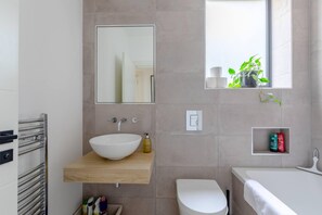 A modern bathroom with a large white sink, a mirror, a toilet, and a bathtub. There is a window with a view of greenery outside.