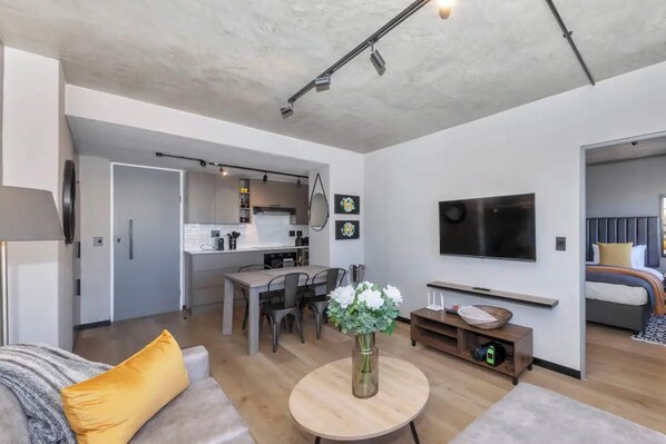 Una sala moderna con un sofá, una mesa de centro y un televisor. Se observa un comedor con una mesa y sillas, y una cocina al fondo.