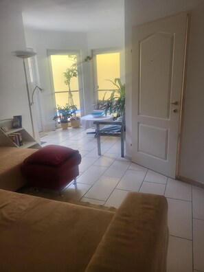 A living room with a sofa, a red cushion, a table with a bowl, and a door leading to a balcony with plants.