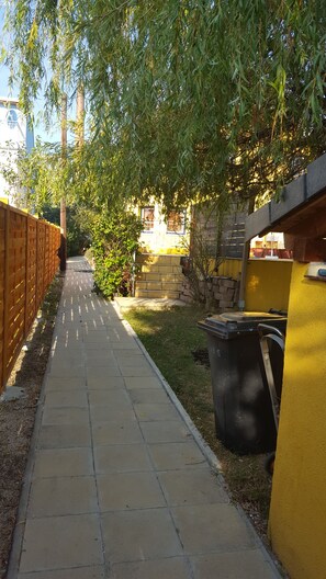 A paved pathway in a courtyard with a wooden fence on one side, a yellow wall on the other, and a black trash bin.