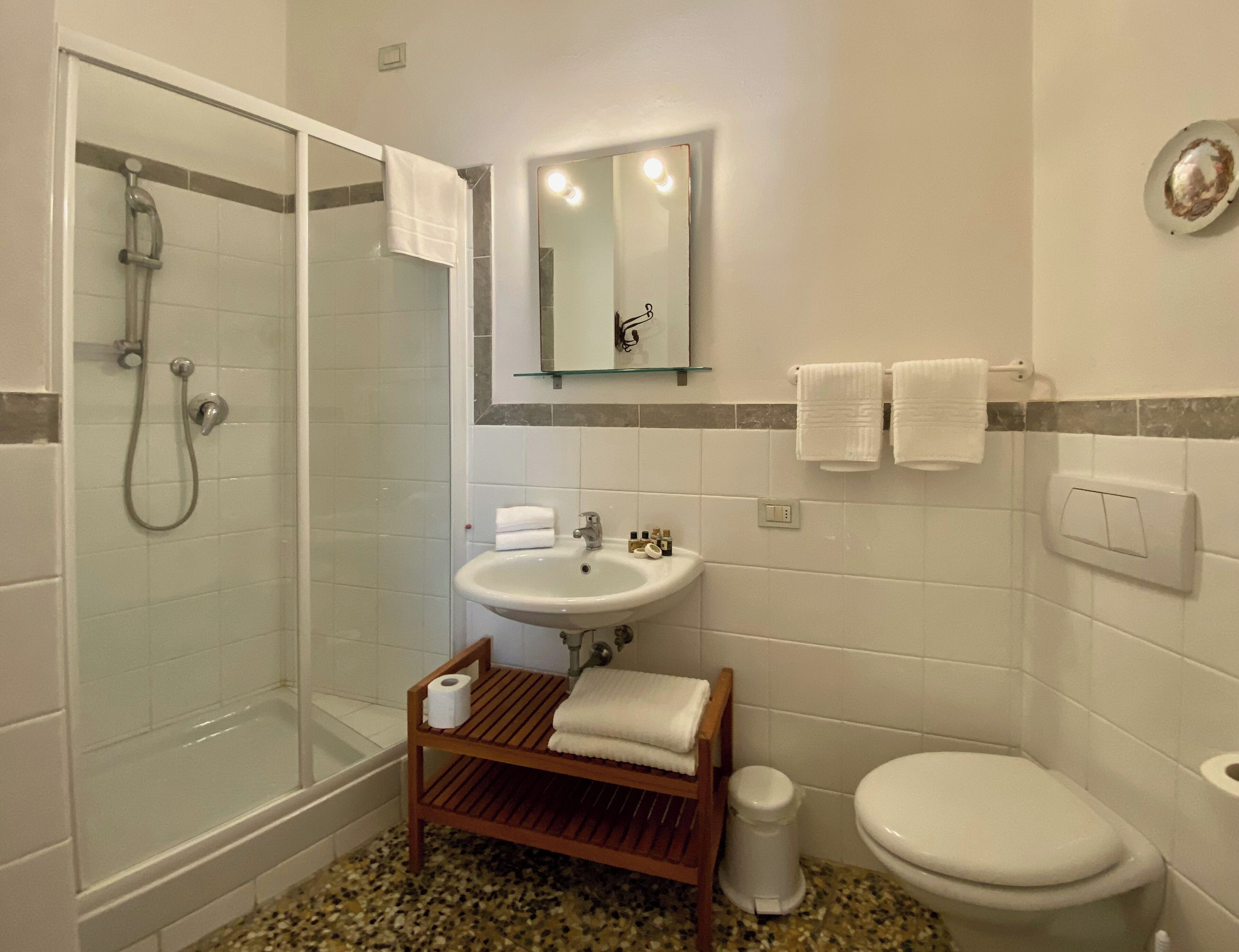 A bathroom with a glass shower enclosure, a sink, a toilet, and a wooden shelf with a towel.