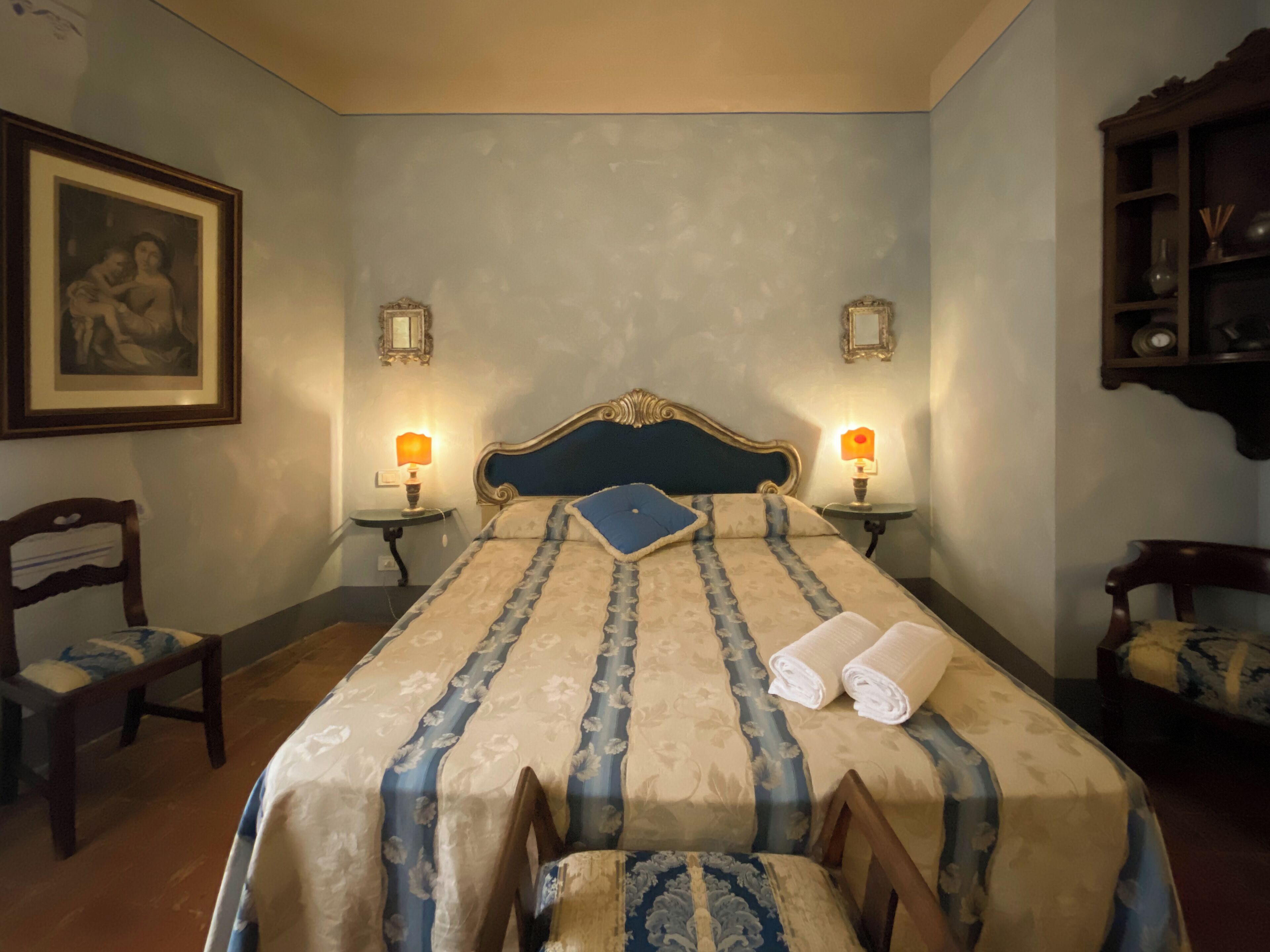 Una cama bien hecha con una colcha estampada, dos lámparas de noche, un cuadro en la pared y una mesita con una silla.