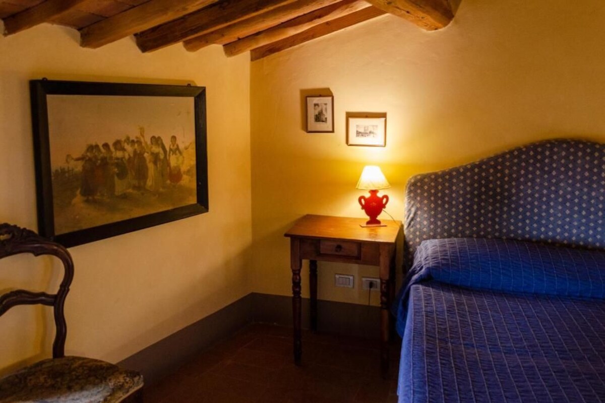 A room with a bed, a wooden chair, a table with a lamp, and a framed picture on the wall.