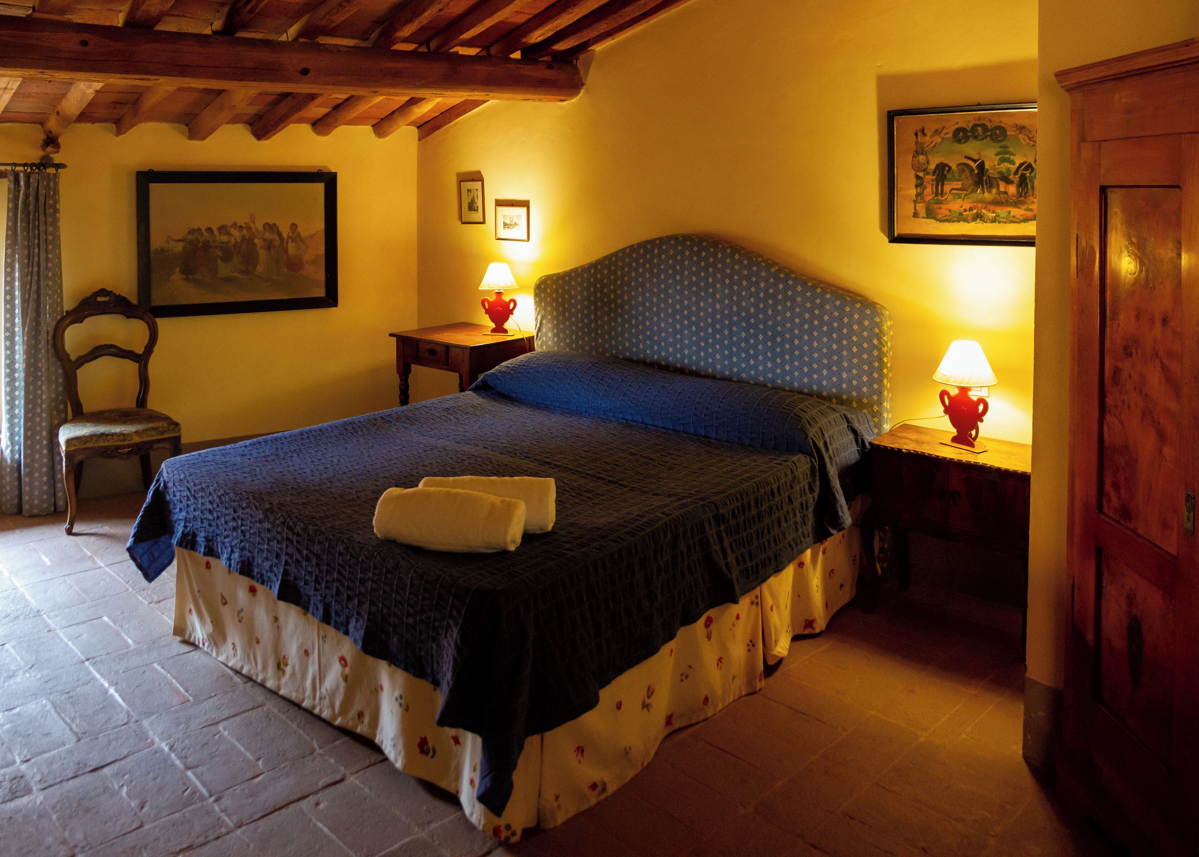 A bedroom with a bed, two bedside lamps, a chair, a wooden cabinet, and a painting on the wall.