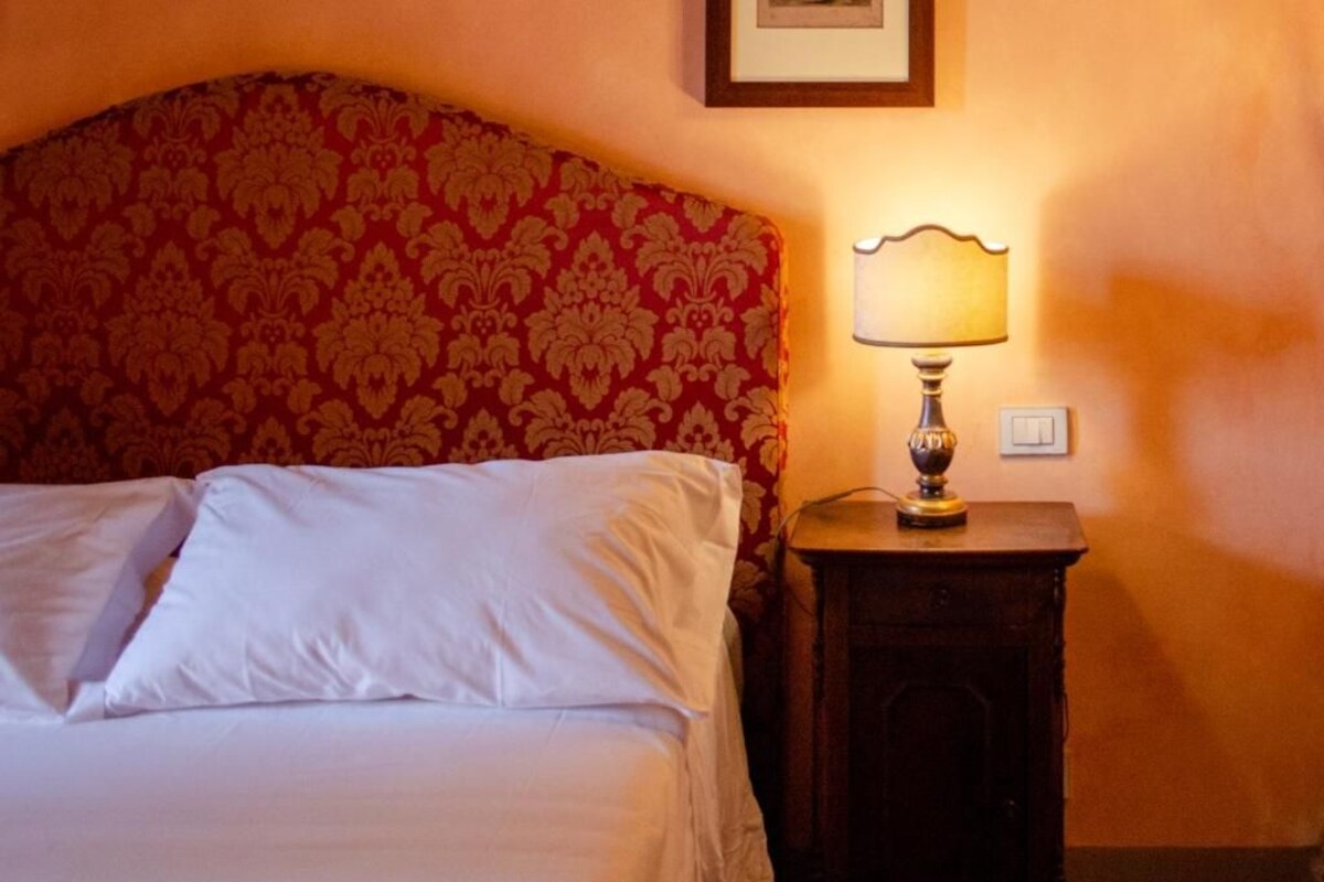 A bed with white linens and a red patterned headboard, a bedside table with a lamp, and a framed picture on the wall.