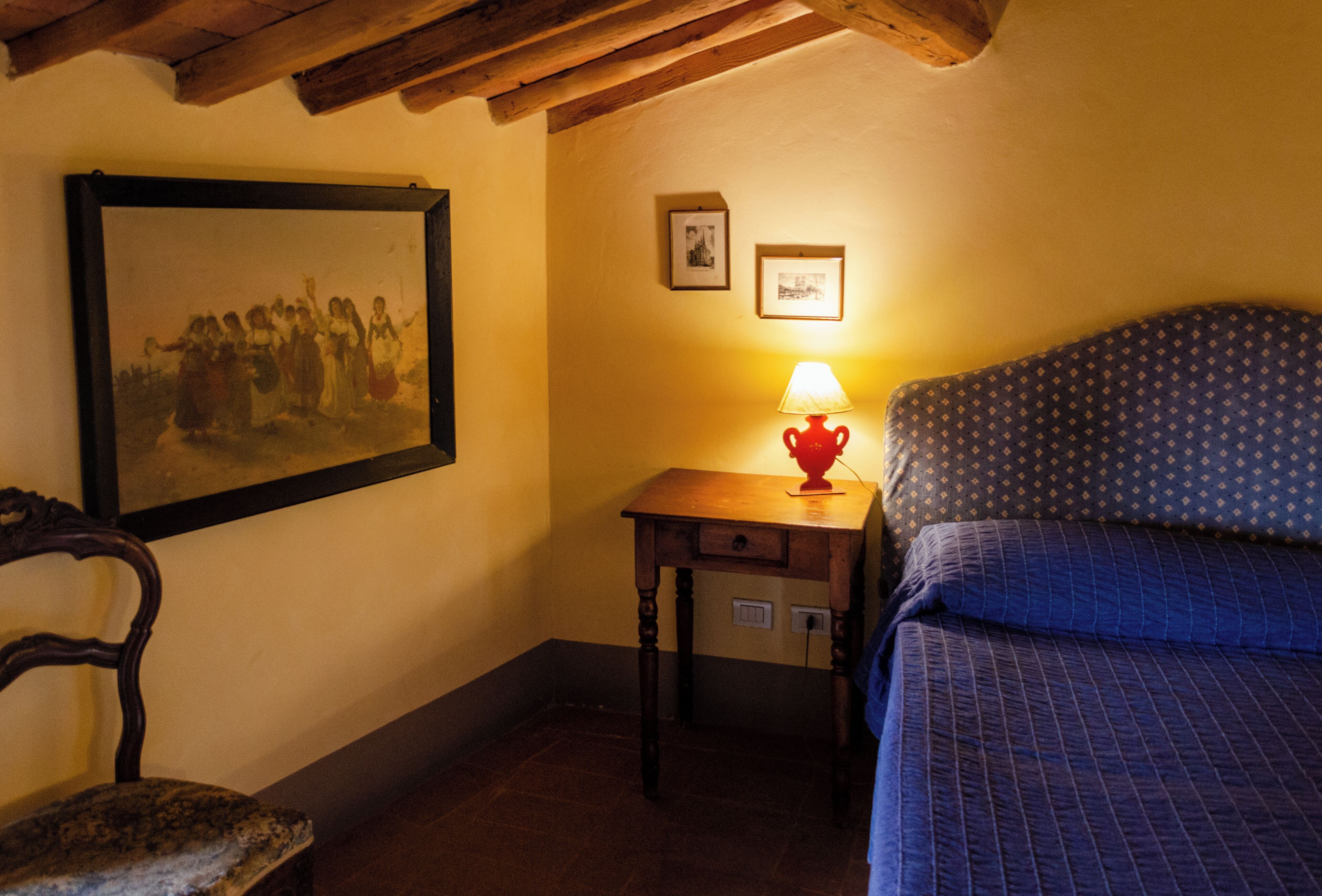 A room with a bed, a wooden chair, a small table with a lamp, and two framed pictures on the wall.