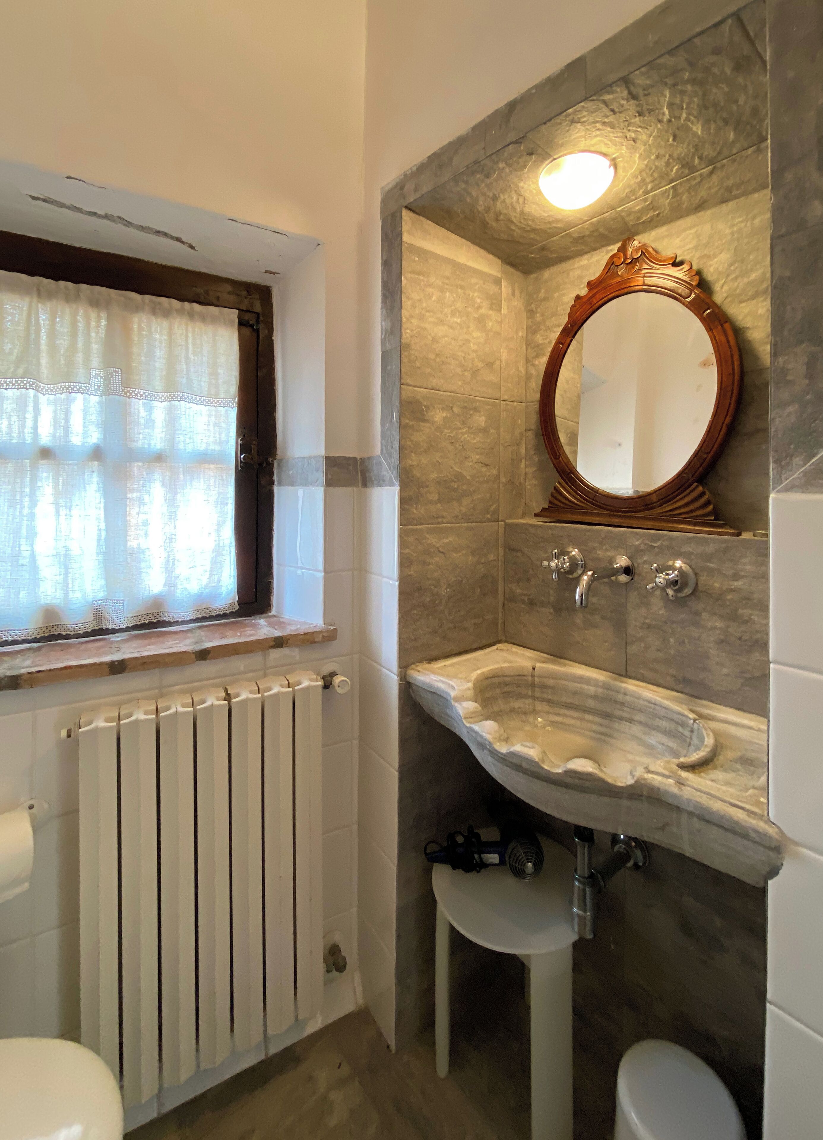 Un baño con un radiador blanco, un lavabo con encimera de mármol, un espejo con marco de madera y una ventana con cortina de encaje.