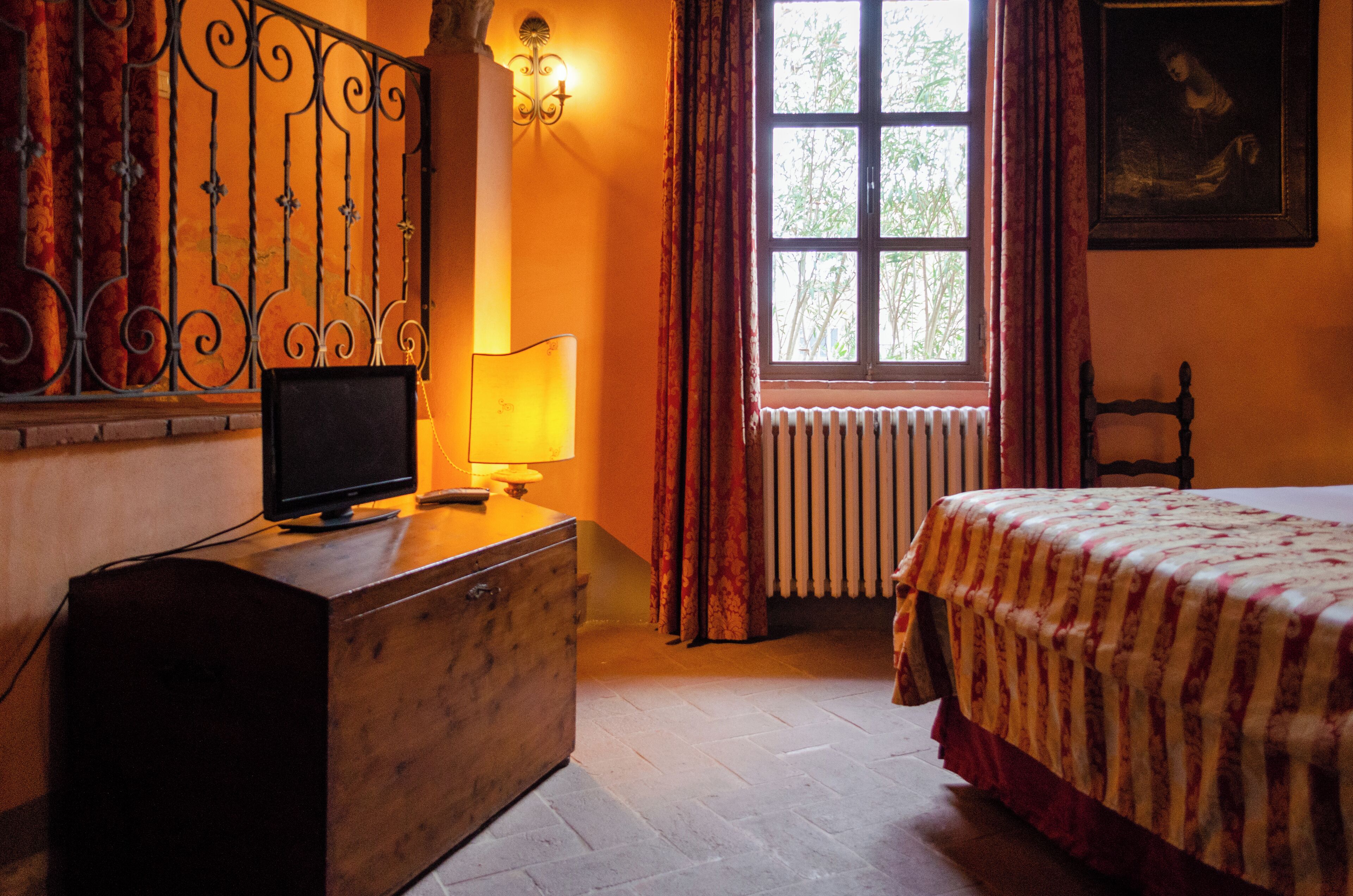 Un dormitorio con cama, cómoda, televisor, ventana con cortinas, radiador y un cuadro en la pared.