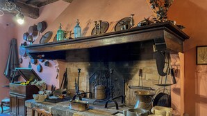A rustic kitchen with a stone fireplace, wooden shelves with kitchen utensils, and a dining table with chairs.
