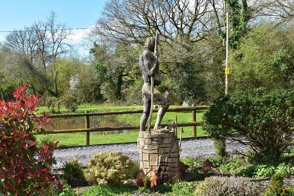 Eine Statue einer Person, die eine Stange hält, umgeben von Grünpflanzen und einem Teich.