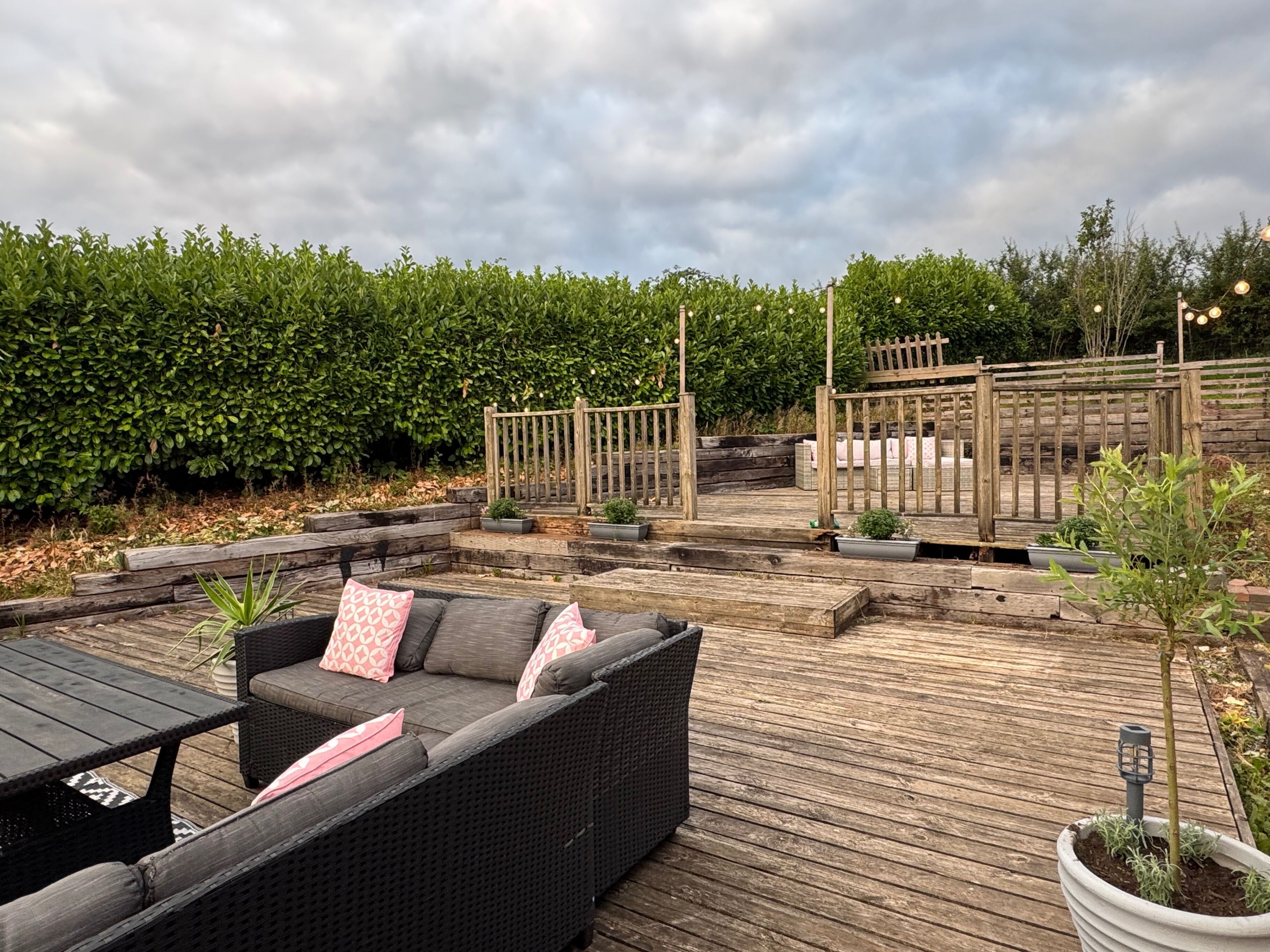 Een houten terras met tuinmeubilair, een houten schutting en een haag op de achtergrond.