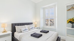 A bedroom with a bed, two bedside tables, a window with blinds, a wardrobe, and a vase with flowers.