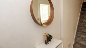 Un pasillo con pared blanca, un espejo ovalado con marco de madera y una pequeña consola blanca decorada con adornos.