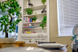 A dining area with a table set for two, a bookshelf with various items, and a window with blinds. - Two-Bedroom Townhouse (Vale of Glamorgan)