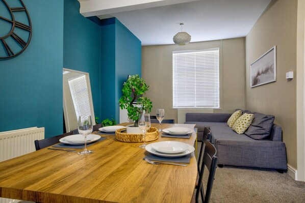 Un coin repas avec une table en bois dressée pour quatre personnes, un canapé gris, une plante en pot et une horloge murale.