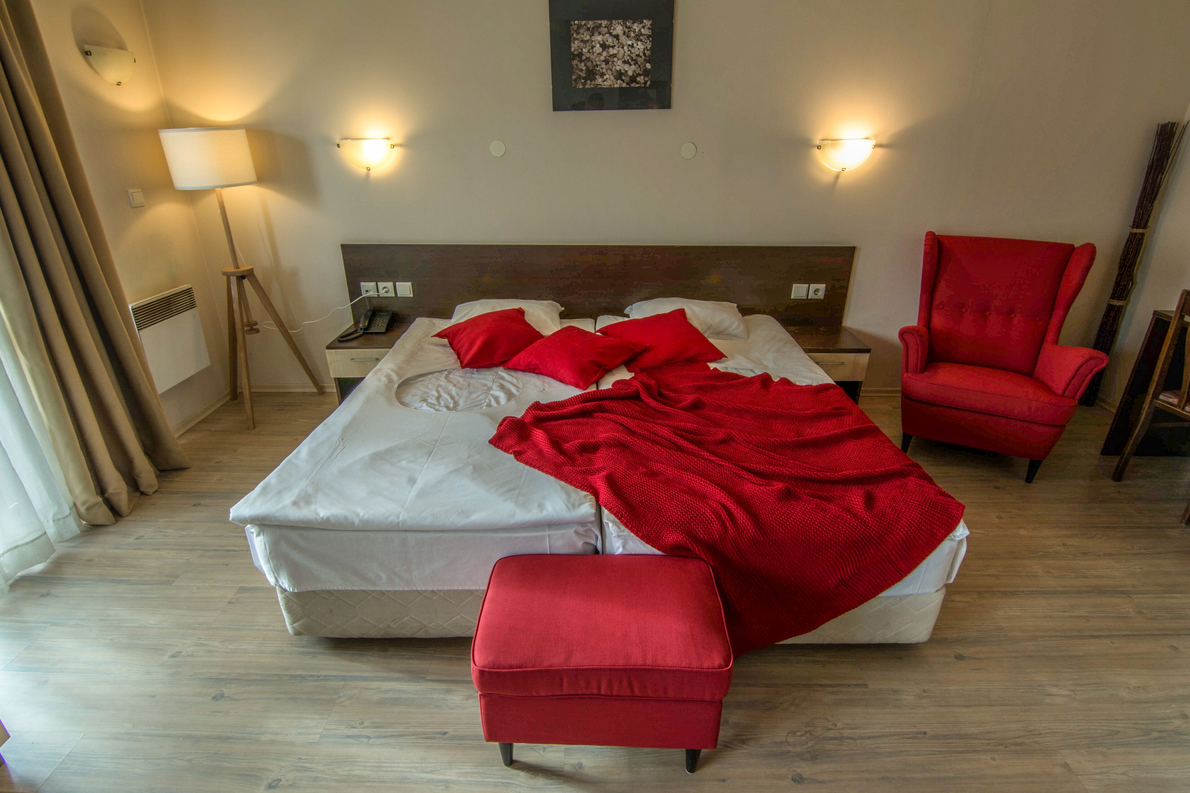 A hotel room with a bed, a red armchair, a red ottoman, a bedside table with a lamp, and a wall-mounted phone.