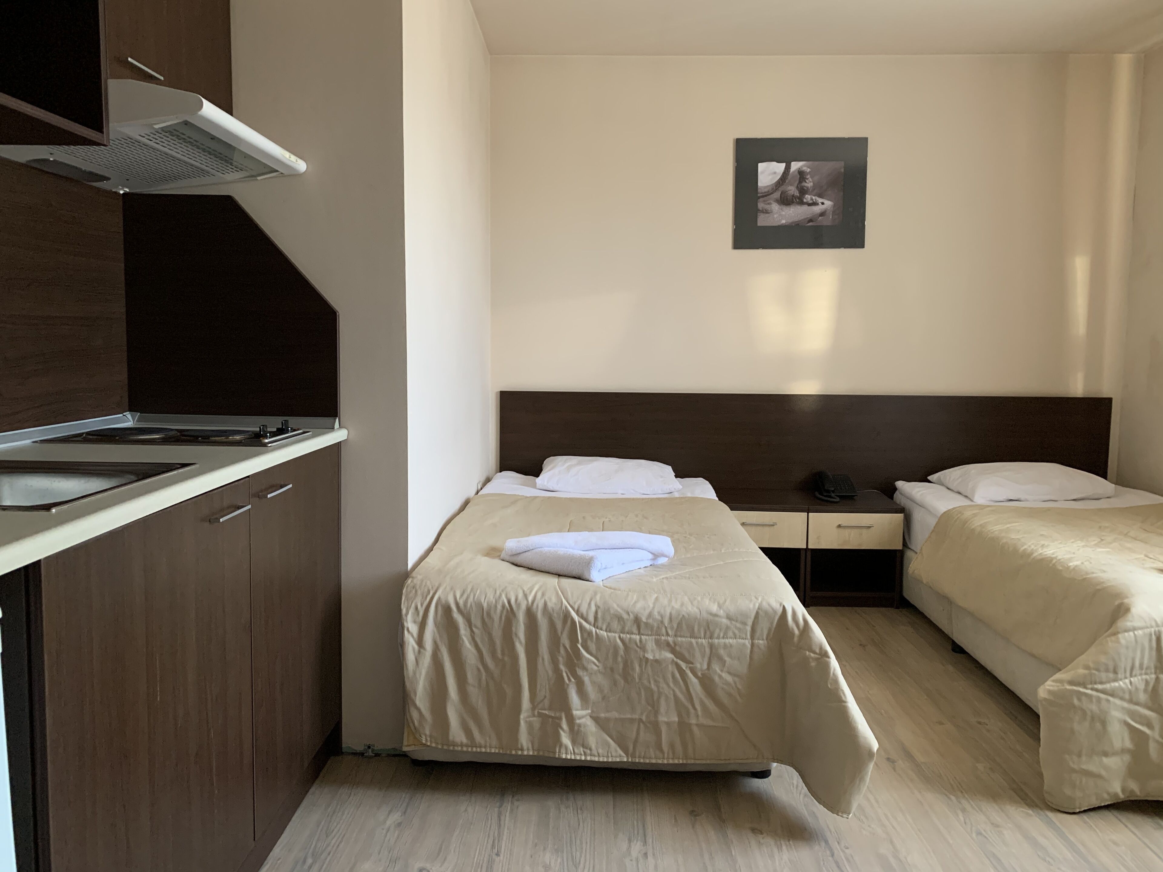 A compact hotel room with two single beds, a small kitchenette, and a framed picture on the wall.