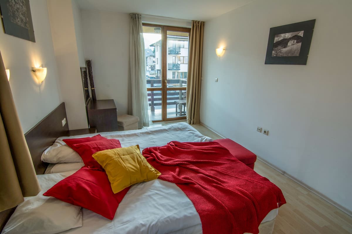 A hotel room with a bed, a red and white blanket, a yellow pillow, and a view of a balcony with a building in the background.