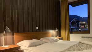 Una cama bien hecha con sábanas blancas, cabecera de madera y una manta decorativa. Hay dos lámparas de noche y una mesita de noche con una lámpara. Se ve una ventana con vistas a un edificio y montañas.
