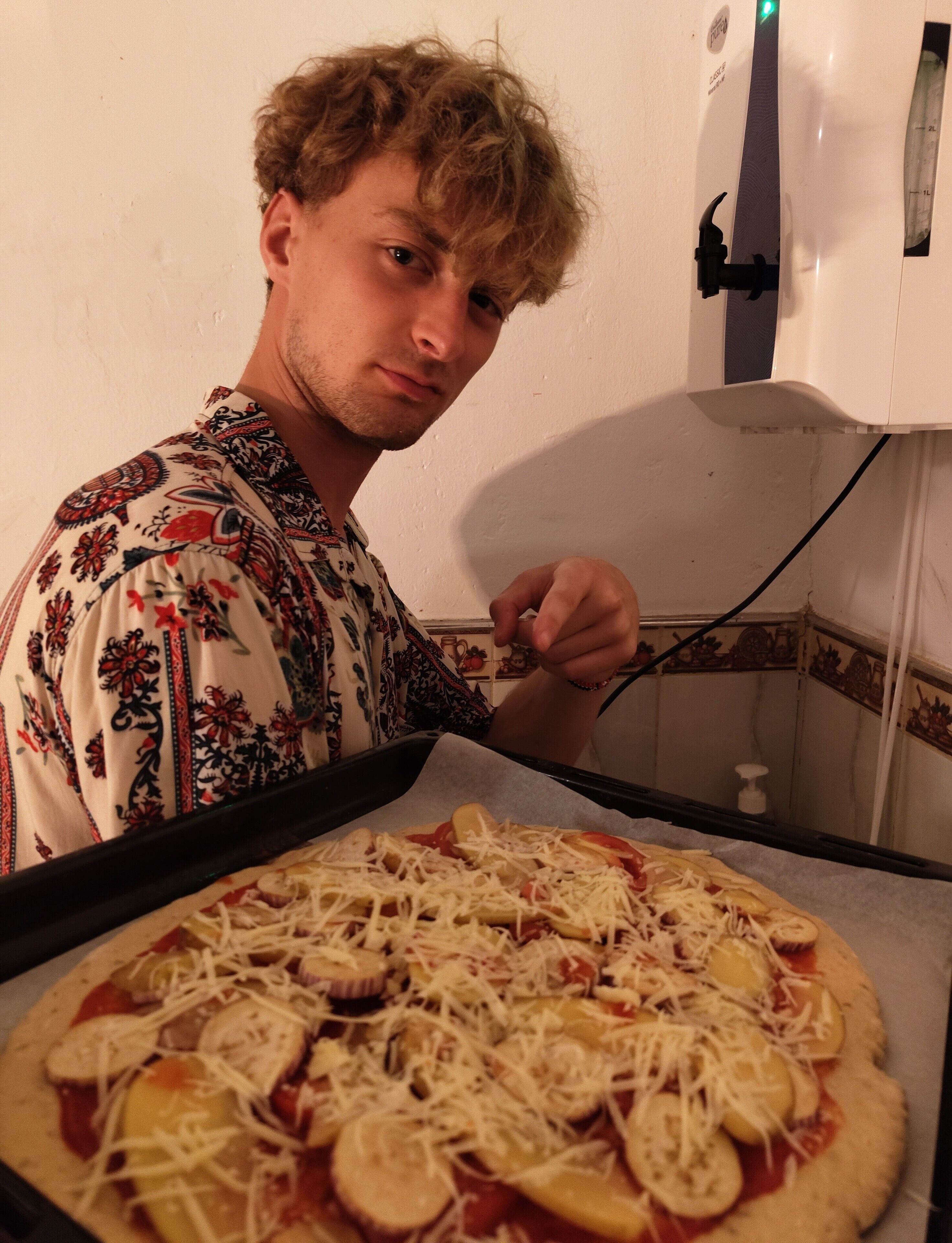 En person i en mönstrad skjorta som förbereder en pizza med ost och grönsaker på en bakplåt.