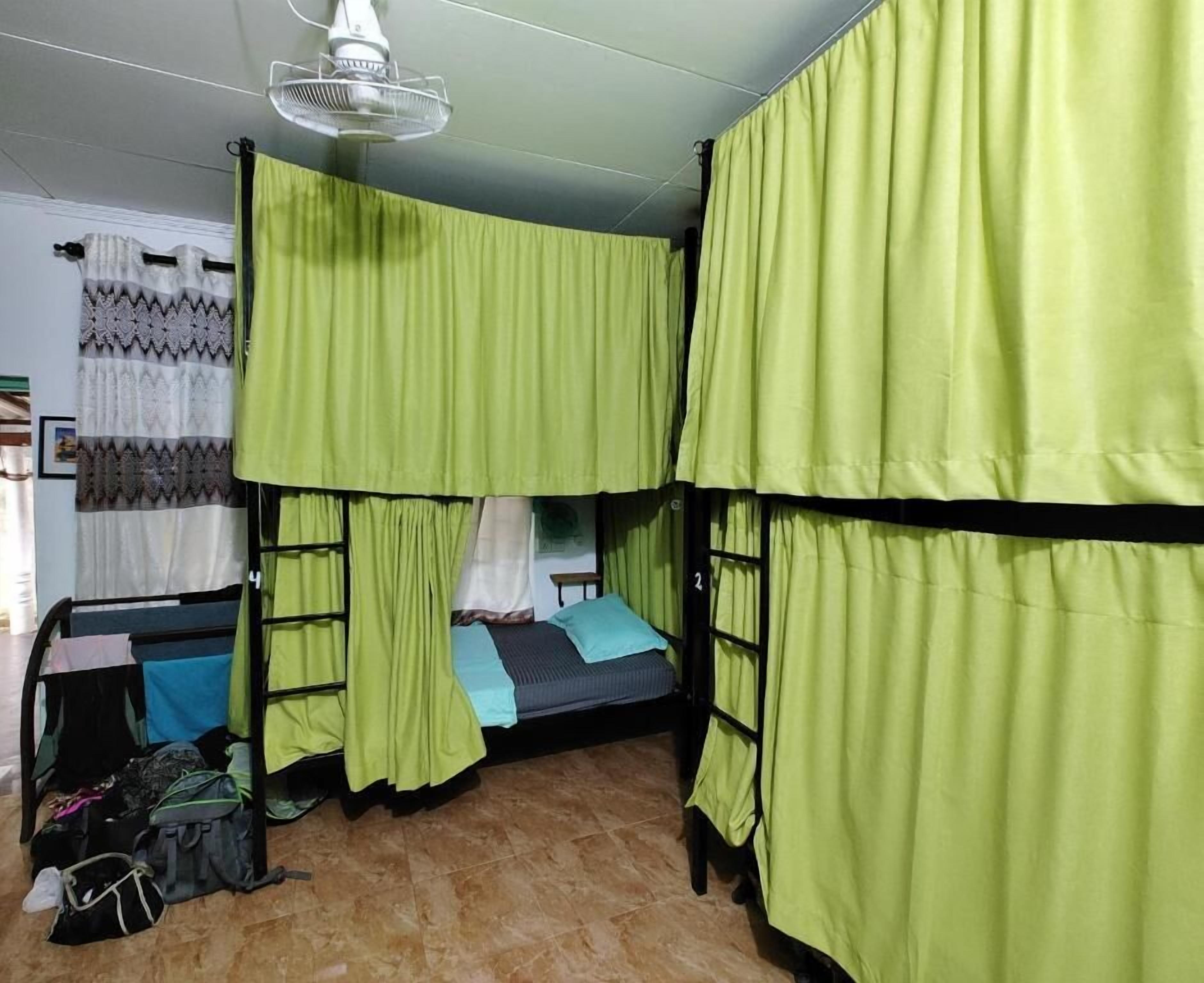 Ein Schlafsaalzimmer mit Etagenbetten, grünen Vorhängen, einem Ventilator und einem Fliesenboden.