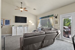 A living room with a large sectional sofa, a ceiling fan, a flat-screen TV, a kitchen area with white cabinets, and a window with blinds.