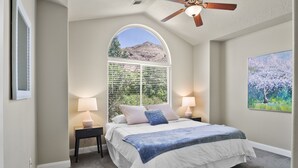 A bedroom with a large bed, two bedside tables with lamps, a ceiling fan, a large window with a view of a mountain, and a painting on the wall. - Perfect location close to Zion! (La Verkin)