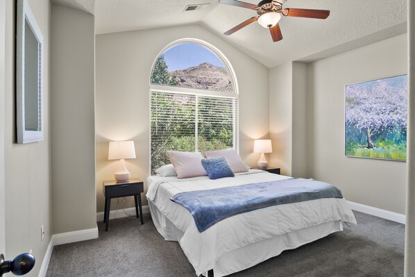 A bedroom with a large bed, two bedside tables with lamps, a ceiling fan, a large window with a view of a mountain, and a painting on the wall.