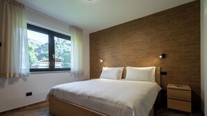 A modern bedroom with a wooden headboard, a large window with curtains, a bedside table, and a nightstand.
