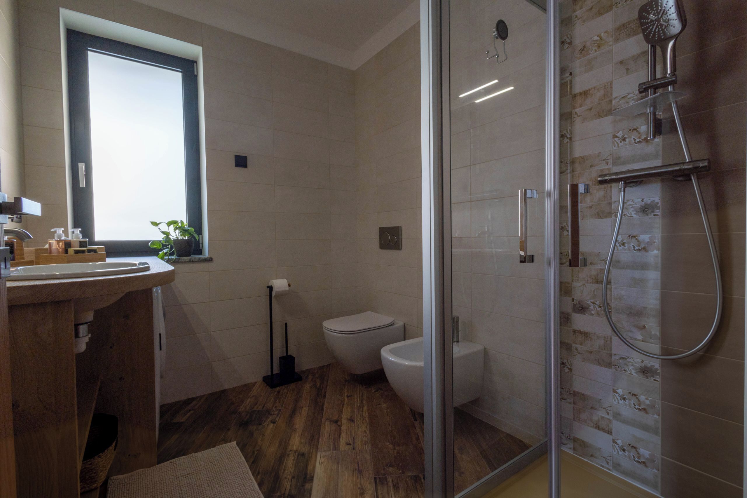Een moderne badkamer met een houten vloer, een douchecabine van glas, een toilet en een wastafel met een raam erboven.