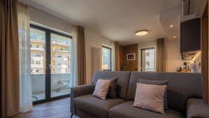 A modern living room with a grey sofa, a sliding glass door to a balcony, and a small kitchen area with a stove and cabinets.