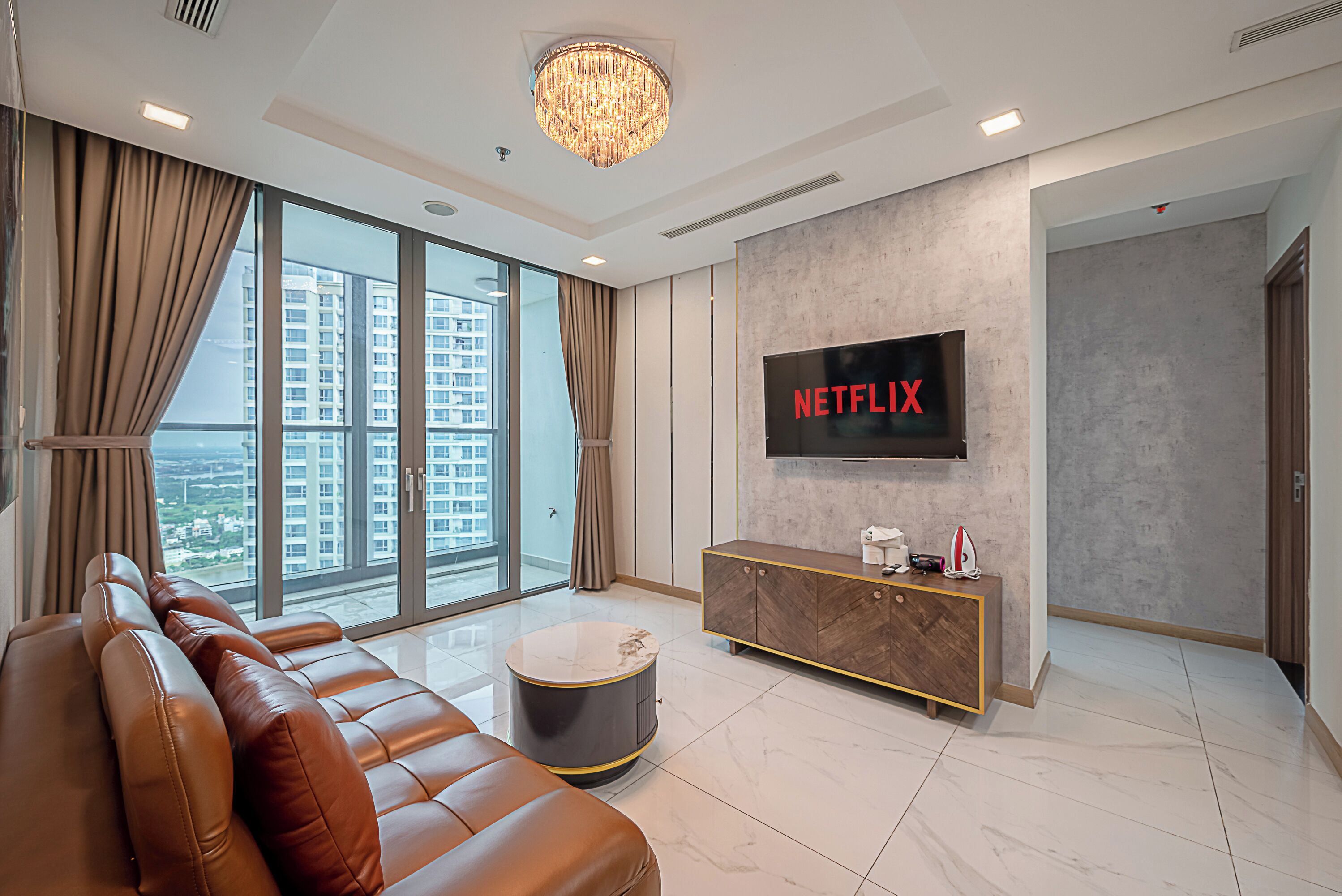 Una sala moderna con un sofá de cuero, una mesa de centro redonda, un televisor montado en la pared y vistas a la ciudad a través de amplios ventanales.