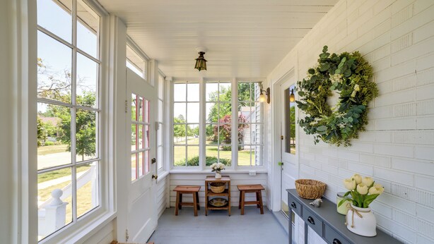 En ljus och luftig veranda med stora fönster, en flätad bänk, ett litet bord med korgar och ett grått konsolbord med en krans och blommor.