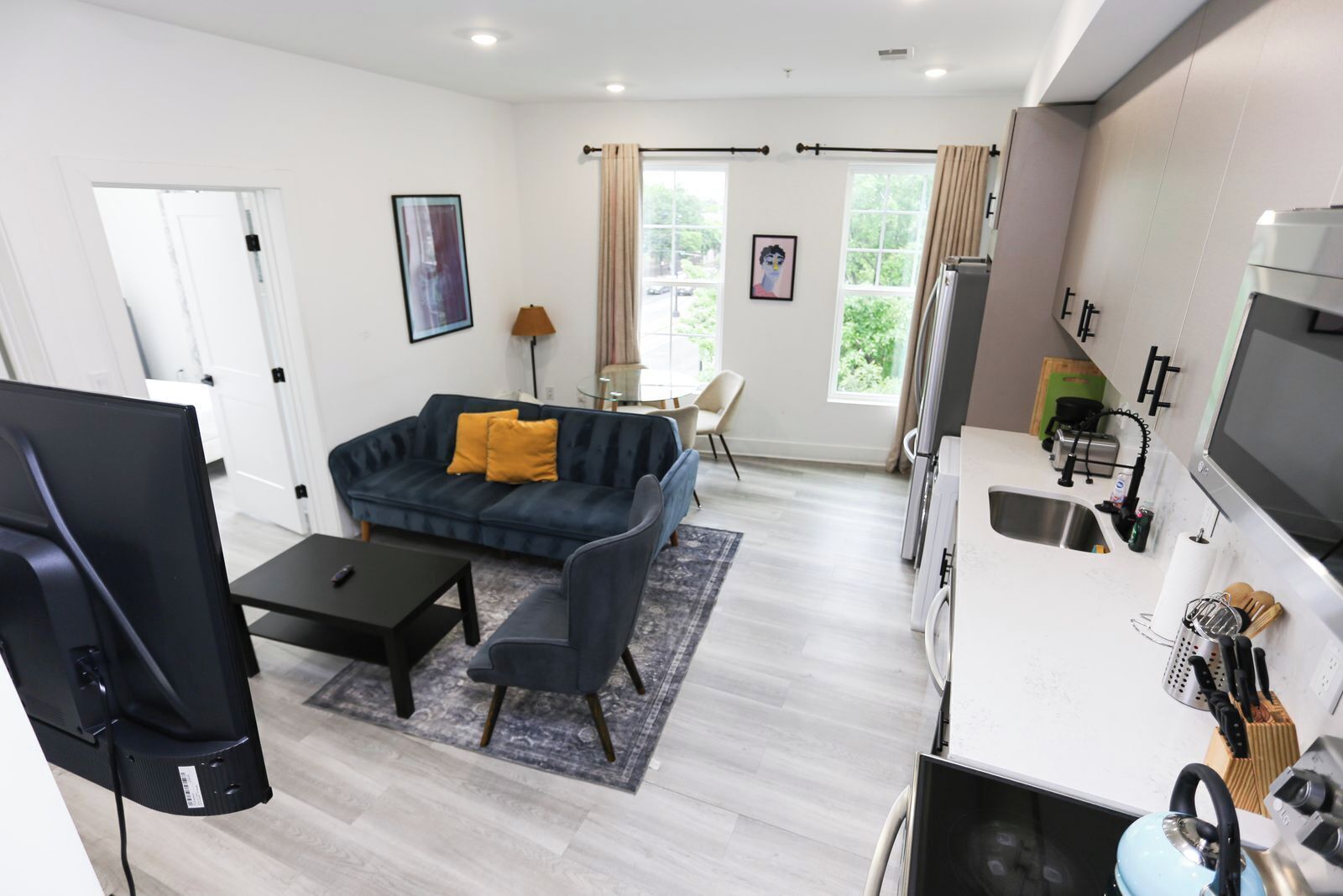 A modern living room with a sofa, coffee table, and TV. There is a kitchen area with a sink, stove, and refrigerator.